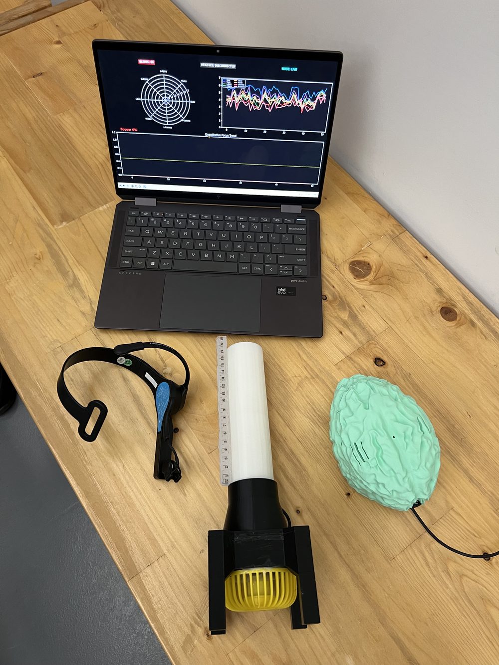 The whole system on a wooden bench: a laptop running the live brainwave dashboard, the EEG headset, the tube on its nozzle with the yellow fan, and the brain-shaped enclosure.