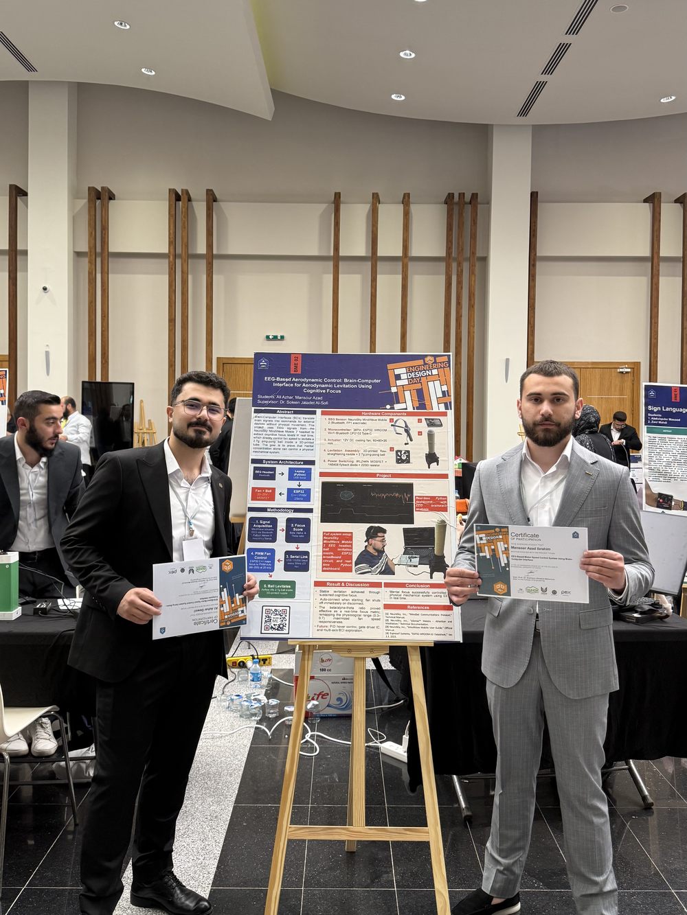 Ali Azhar Dewali and Mansour Azad standing either side of the project poster on an easel, each holding a certificate of participation.