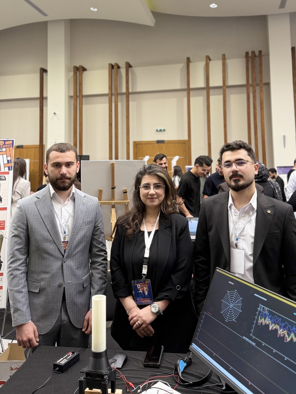 The two students and their supervisor standing behind the project stand, with the tube and the dashboard monitor on the table in front of them.