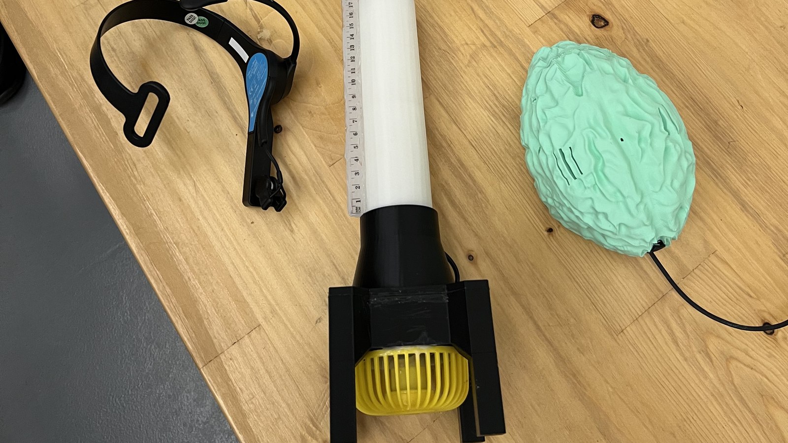 The system laid out on a bench: the EEG headset, the tube on its 3D-printed nozzle with the yellow fan beneath it, and the mint-green brain-shaped enclosure.